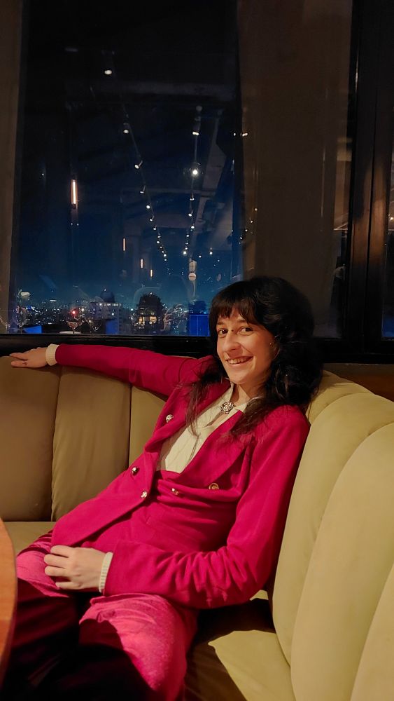 eu (nao binario branco palido de cabelo castanho shaggy) de terninho vermelho sorrindo sentado num banco estofado em frente a uma janela grande com a paisagem da vista de sao paulo a noite ao fundo