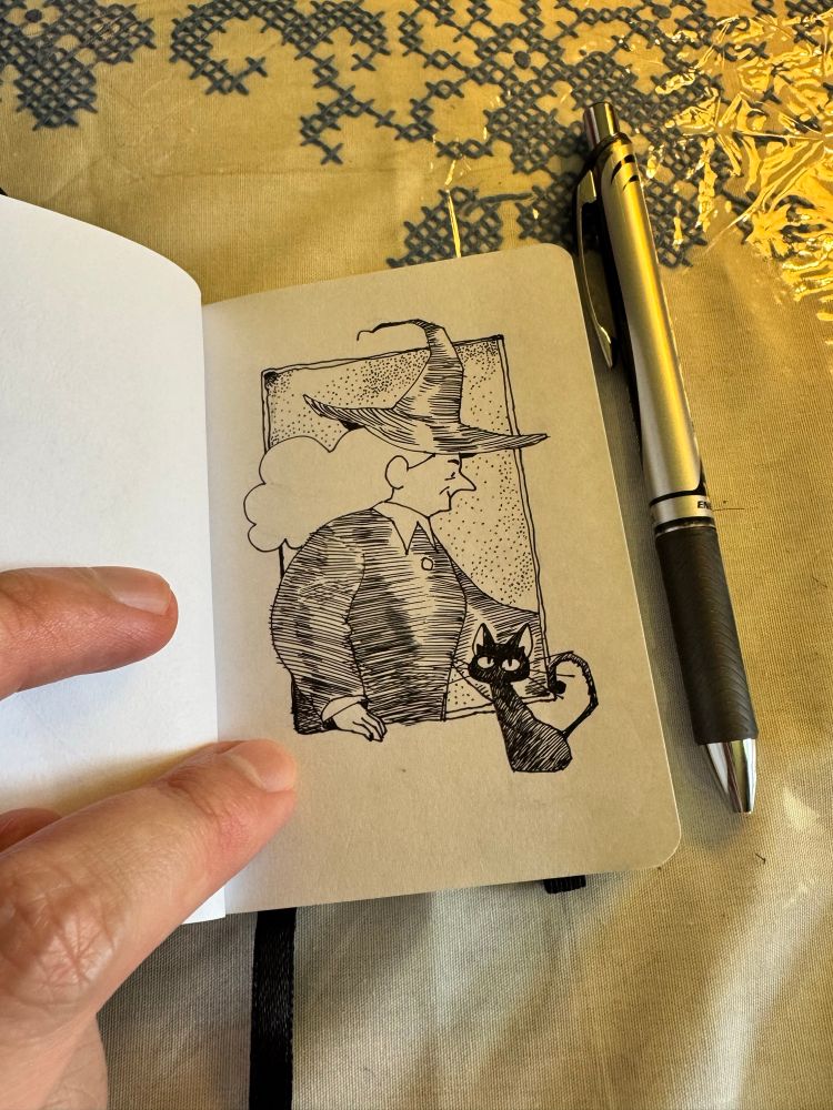 Under a faint yellow light, I hold open a very small sketchbook, which is next to the pen I used inside the sketchbook. There is a small drawing of a witch and her black cat inside it. 