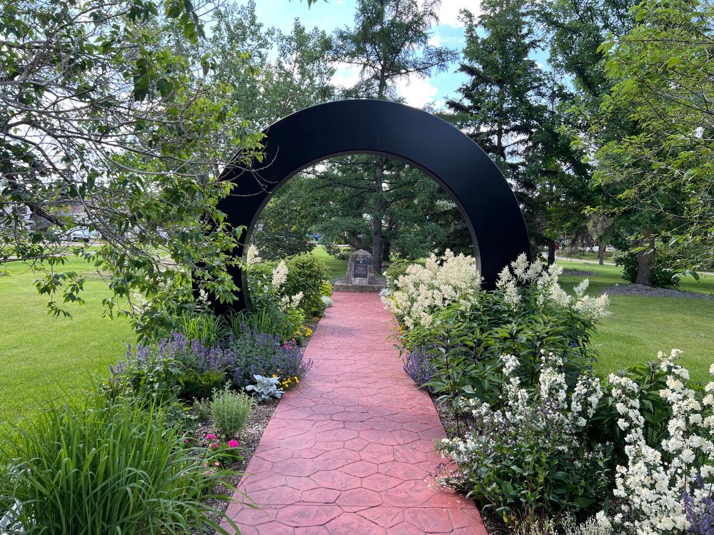 A red stone pathway passes through a circular archway, lined with flowers. A monument is at the end of the path 