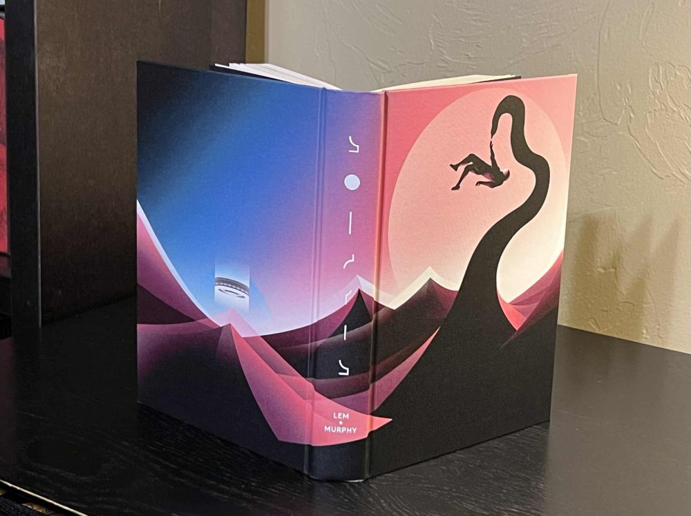 Photo shows the front and back covers as well as the spine of a clothbound edition of the novel Solaris. The design on the cover stars dark at the bottom and then slowly shifts to a pink, finally shifting to blue to show the sky. 