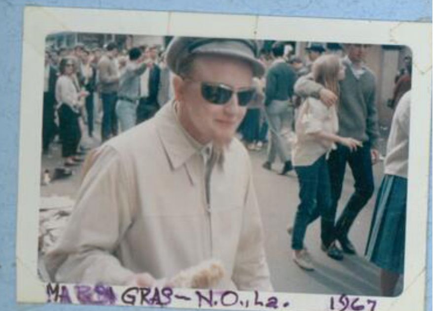 White trans man with a hat and sunglasses hold corn with celebrating people in the background 
