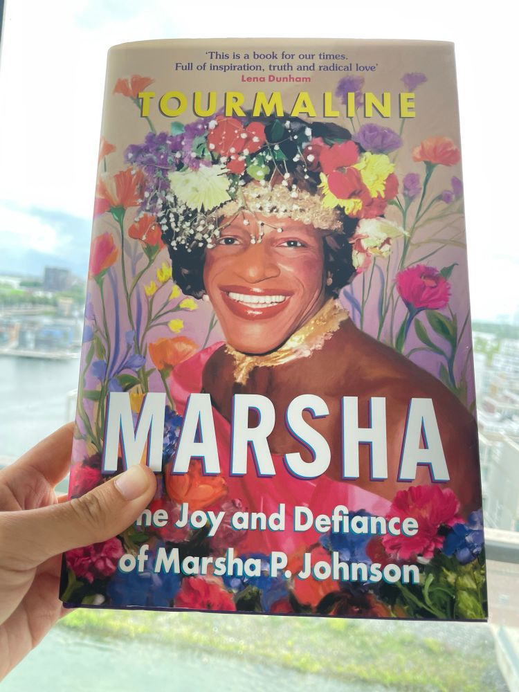 Colorful, flowery, and haptically satisfying book cover featuring an image of Black trans activist. On top blurb by Lena Dunham: 'This is a book for our times. Full of inspiration, truth and radical love'
Author’s name TOURMALINE below.
Bottom of cover, book title 
MARSHA: The Joy and Defiance of Marsha-P, Johnson
(My hand holding the cover is blocking the T)