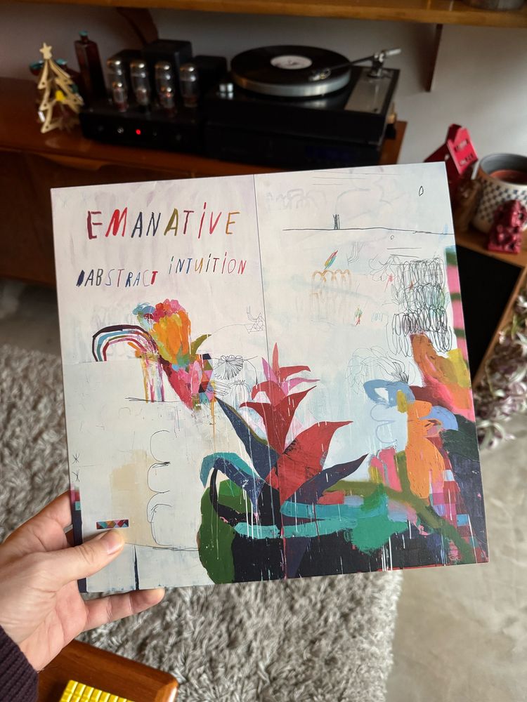 Holding a vinyl copy of the album Abstract Intuition by Emanative in front of a record deck next to a valve amplifier. Album artwork is a painting by the artist in a child-like style with rainbow colours and what look like flowering plants. 