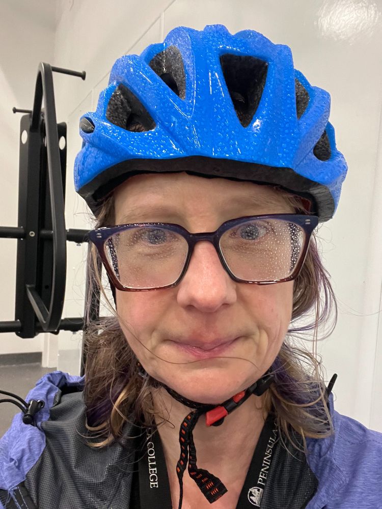 Middle aged white lady wearing a bike helmet and glasses beaded with water inside a bike garage