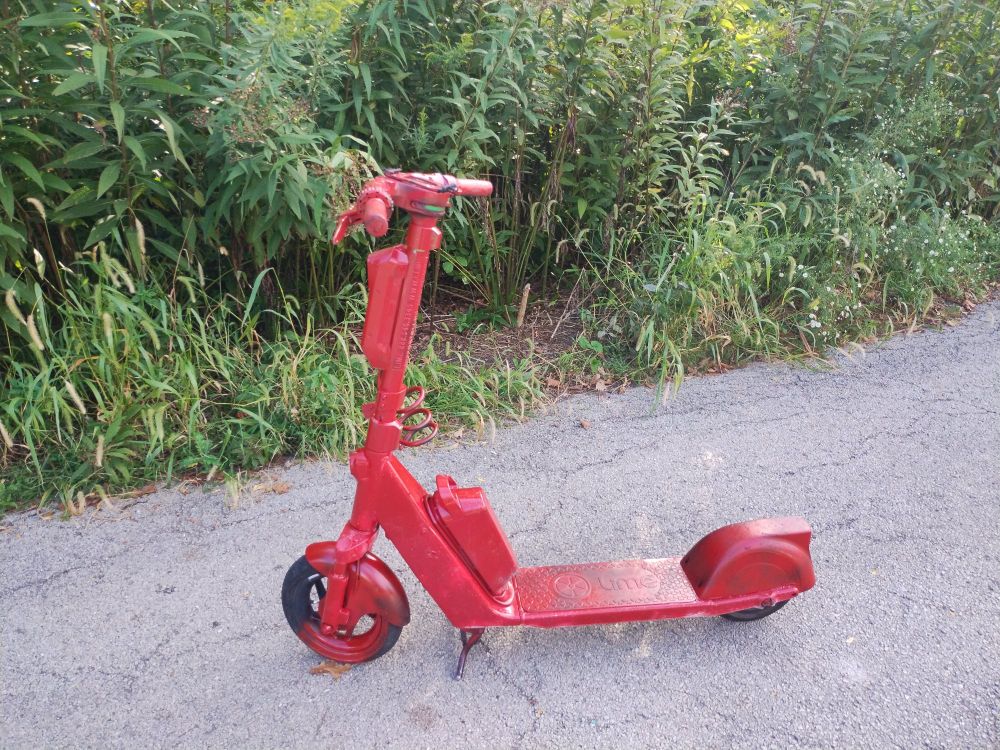 A Lime scooter is sitting on a walking path, coated in red paint. The paint is uneven on the wheels, so it looks like a bad spray paint job.