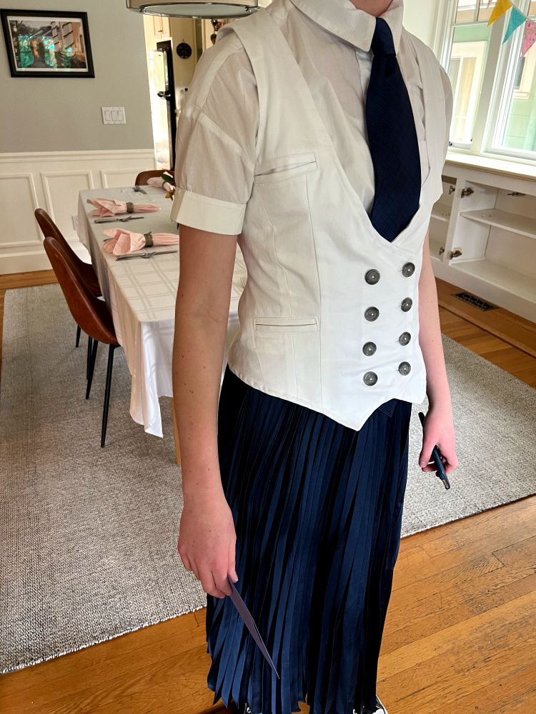 Photo of a white woman standing in a dining room wearing a white shirt and black tie under a hand-made white vest/waistcoat. The double-breasted vest has two columns of dark buttons and three small pockets. The woman is also wearing a long, navy pleated skirt. 