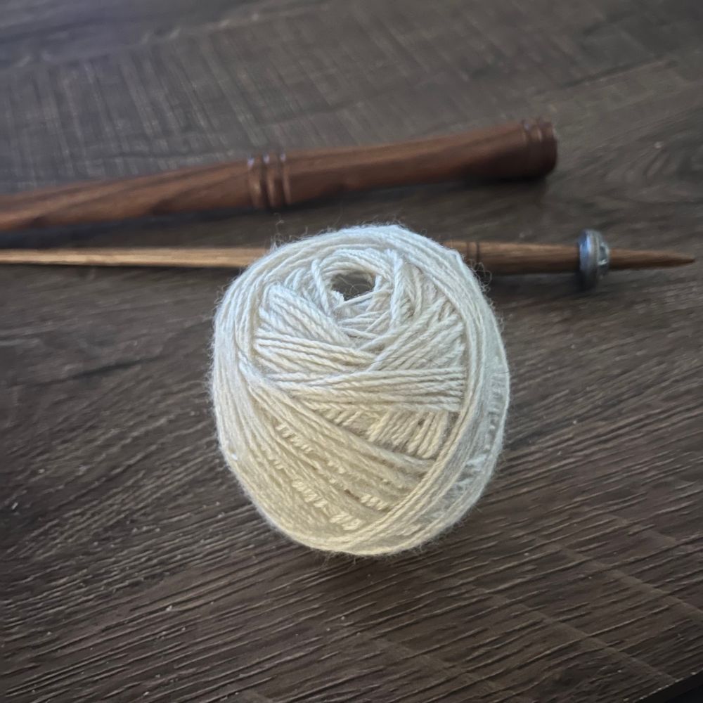 A picture of a hand wound ball of yarn sits on a table. The yarn is cream coloured. In the back ground are a wooden nostepinne and a spindle stick with a pewter whorl 