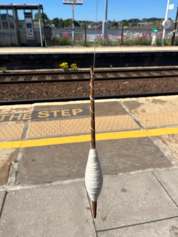 A spindle stick loaded with a medium sized cop of natural cream coloured yarn hangs in the centre of the picture from a length of spun yarn. In the background, slightly out of focus we can make out railway tracks and scenery