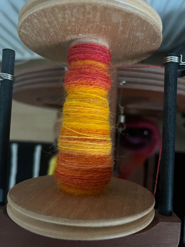 Top down view of a bobbin on a spinning wheel showing an in progress singles yarn. Yellow red and orange colours are visible 