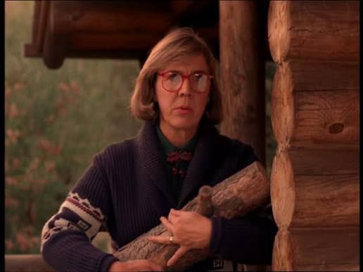 This is the log lady from Twin Peaks. She stands beside a log cabin wall holding a log. She has shoulder length bob hair, and large red glasses. She is wearing a purple knitted cardigan with patterned detail on the sleeve. Behind her is wallpaper featuring plants. She stares concernedly out of the photo 