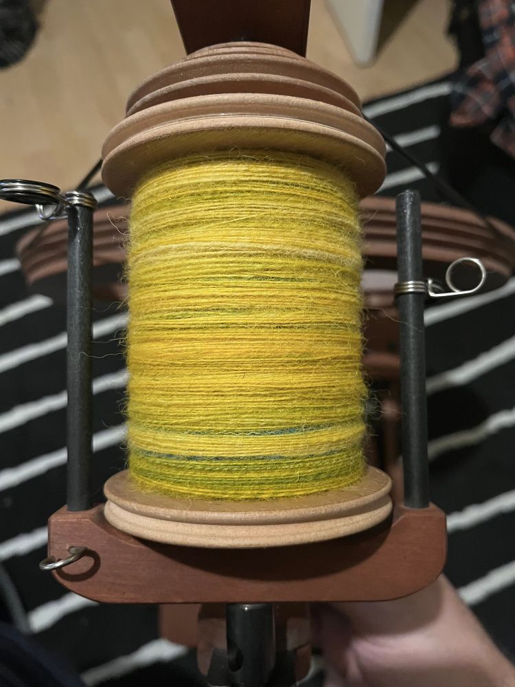 Top down view of a full bobbin on a spinning wheel. The yarn is a thin singles and almost entirely yellow, although you can see green colours poking through from beneath. The bobbin is quite evenly filler, with very little bumping of the yarn at the sides 
