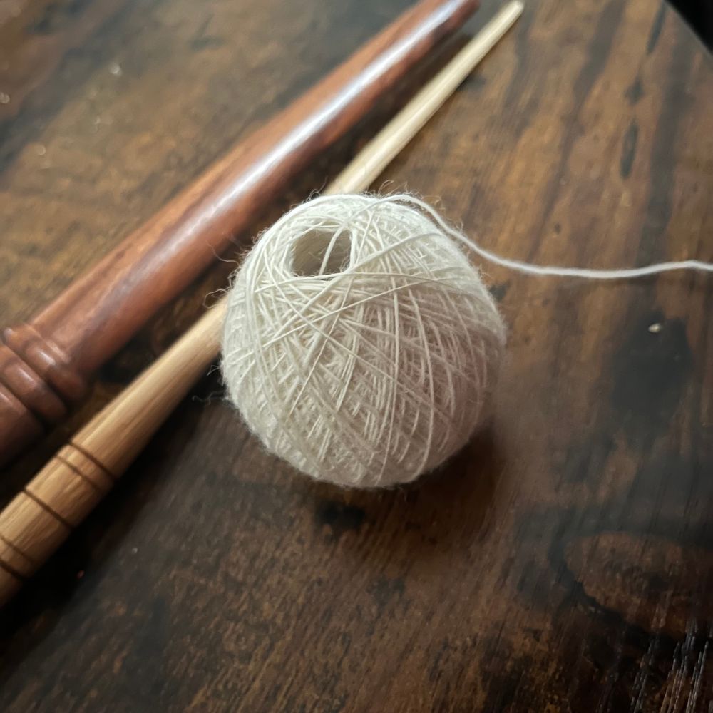 This shows a small hand wound ball of thin singles yarn sitting on a dark wood table. In the background you can see the spindle stick used to spin it and the nostepinne used to wind it 