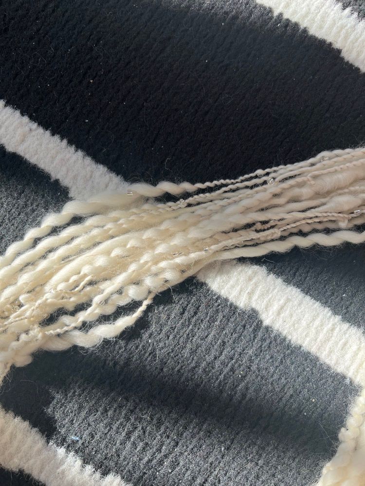 A close up of a section of yarn on a black and white rug. You can see the curly spiral texture of the yarn that moves between thin and thick spirals. Strewn throughout are some small glass seed beads that are plied into the yarn