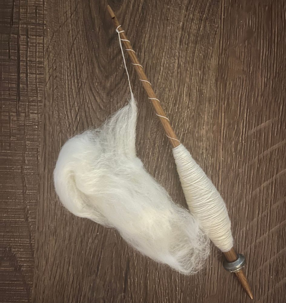 A spindle stick with a pewter whorl lies on a wooden surface. The spindle is loaded with cream coloured thin thread that forms a large cop near the end and then spirals up the spindle. The thread is still connected to a puff of spinning fibre which is visible next to the spindle 