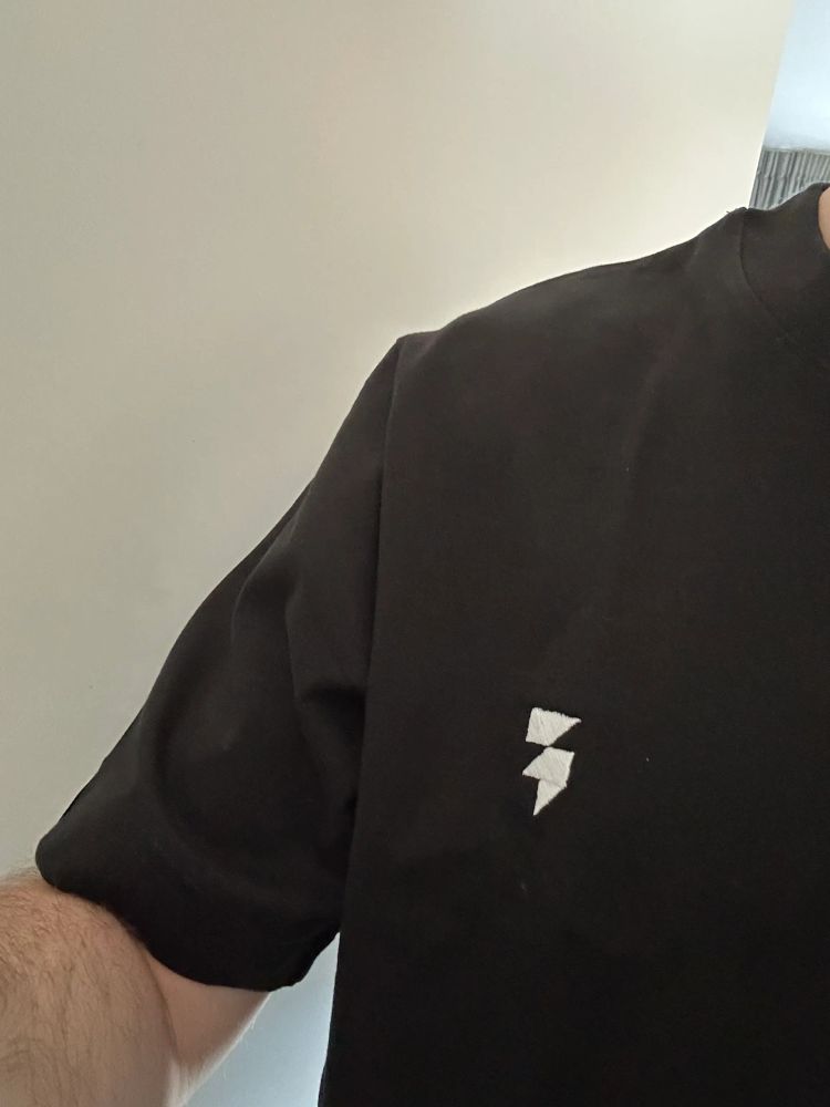 Person's torso wearing a plain black t‑shirt with a small white lightning bolt logo on the chest against a light wall background