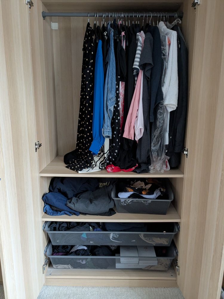 Kleiderschrank bestückt mit Kleidung. Oben hängen Kleider. In der Mitte sind Hosen, Shirts. Darunter Pullover und Sportsachen und da drunter Strumpfhosen, Unterwäsche. 