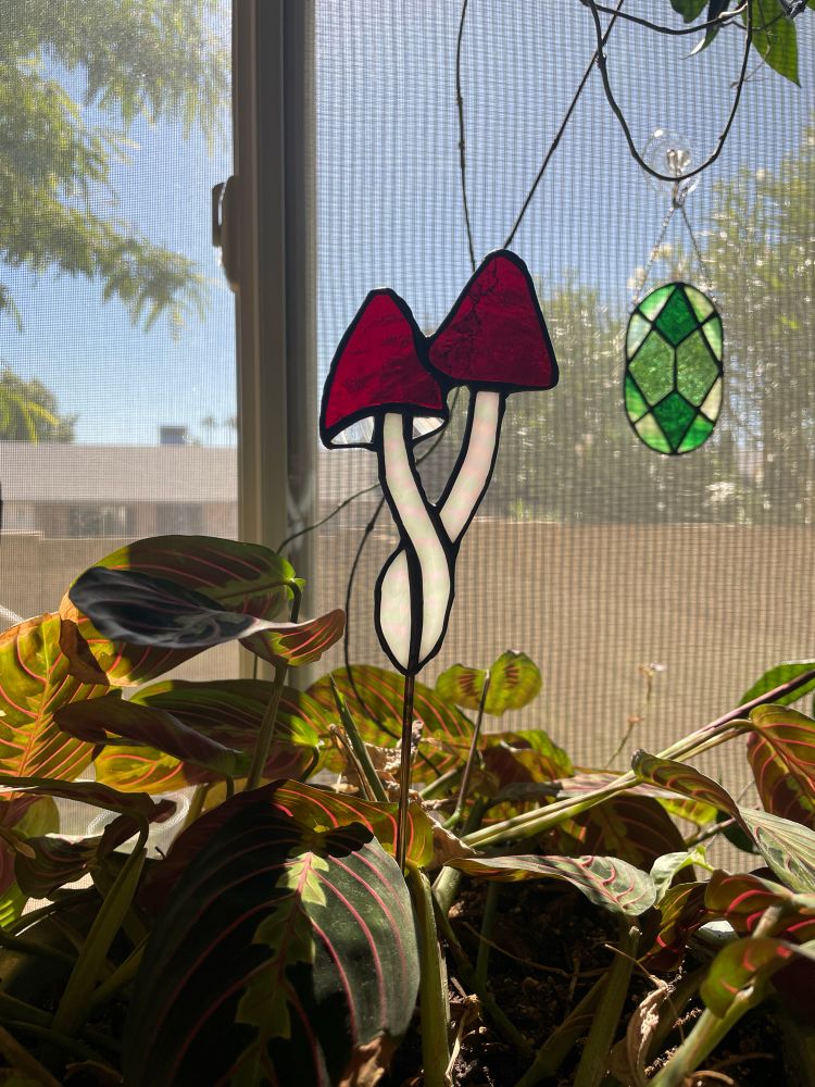 a stained glass plant stake of a cluster of 2 mushrooms. the mushrooms have red caps and white stems. the piece is staked in a houseplant pot 