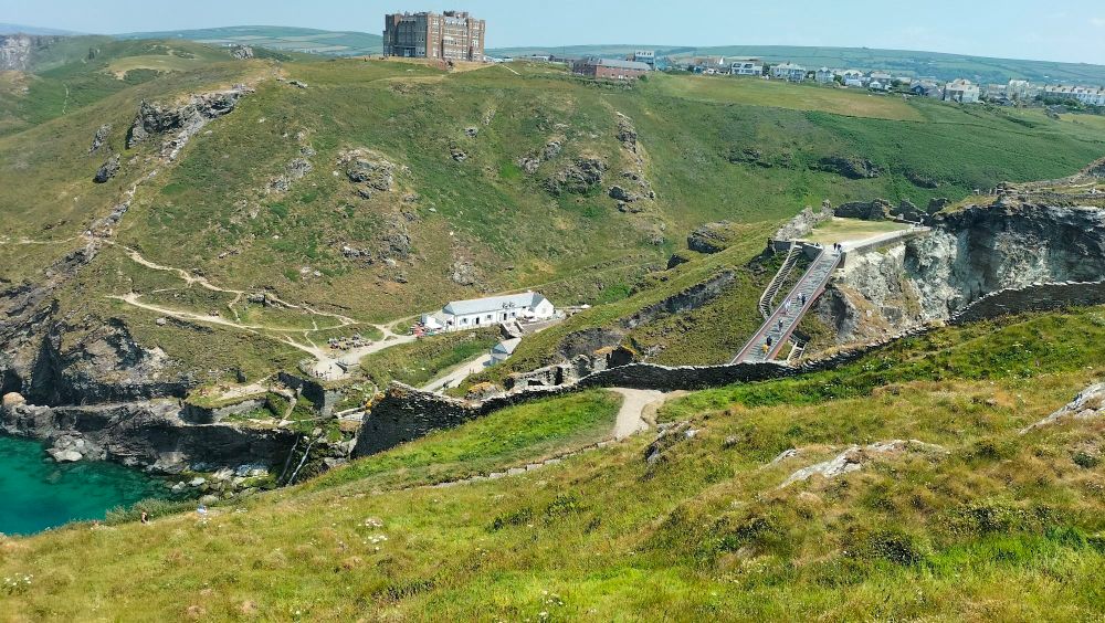 A scenic view taken from within the ruins of Tintagel castle, in Cornwall. The hill I'm on is littered with walls reduced to almost nothing and suspension bridges built to help tourists navigate the ruins. However, on the foot of the hill right in front of me stands the reception, and on the top of that hill another grand ancient cuboidal building stands, still intact.