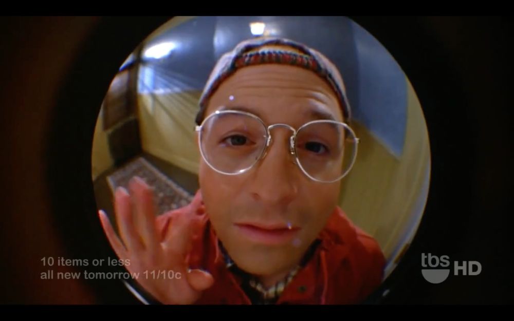 george costanza in an apartment hallway, peering into a front door peephole and waving, with a tragically desperate expression on his face