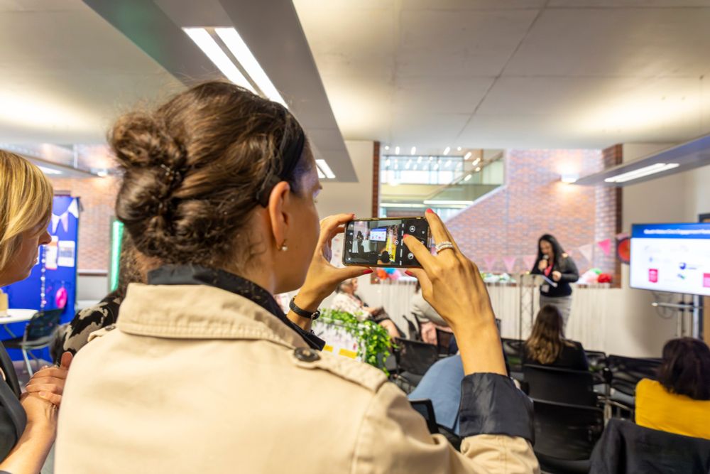 Woman taking a photo on her phone of another woman doing a presentation on a small stage. 
