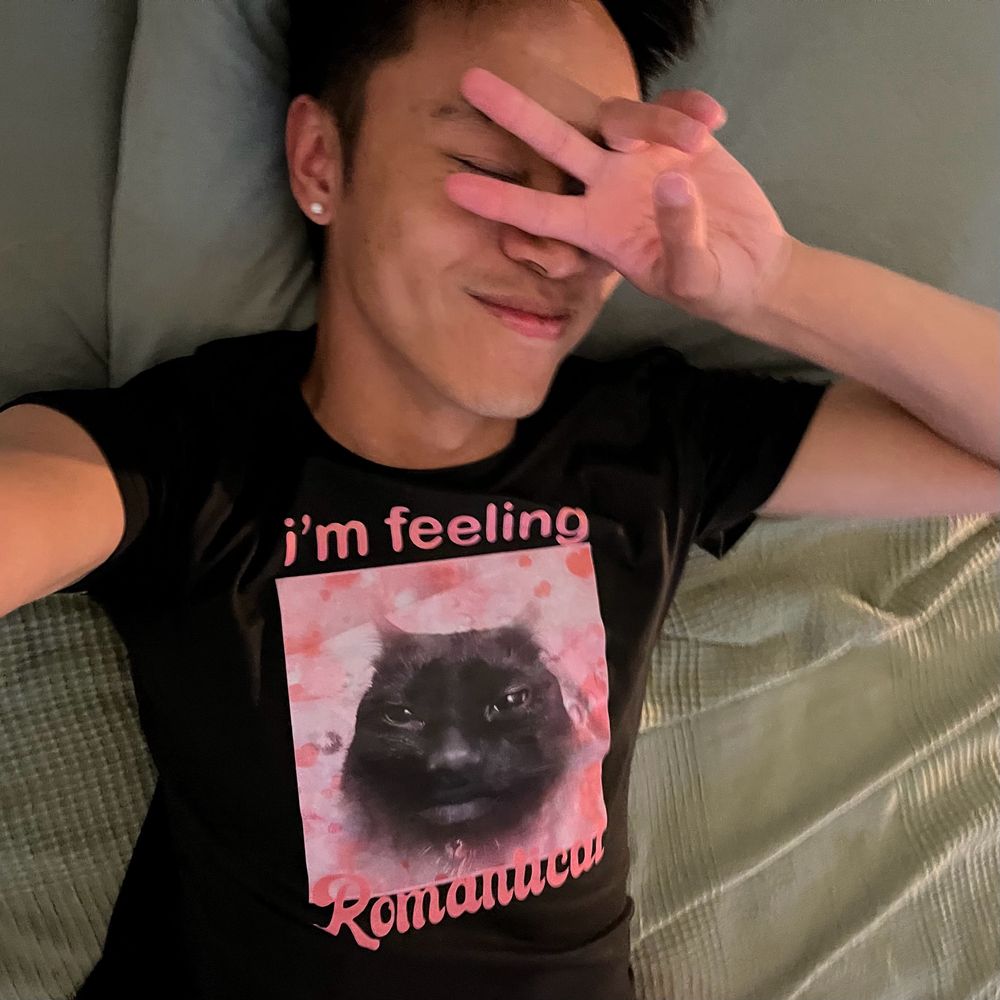 Brandon throwing a peace sign in a black tee featuring a black cat over a pink background that says “i’m feeling romantical”
