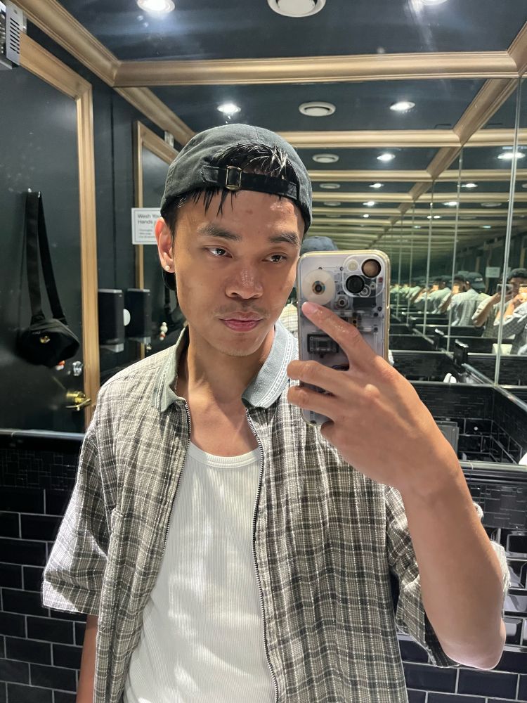 Brandon taking a mirror selfie in a black cap, white tank top and green checkered shirt