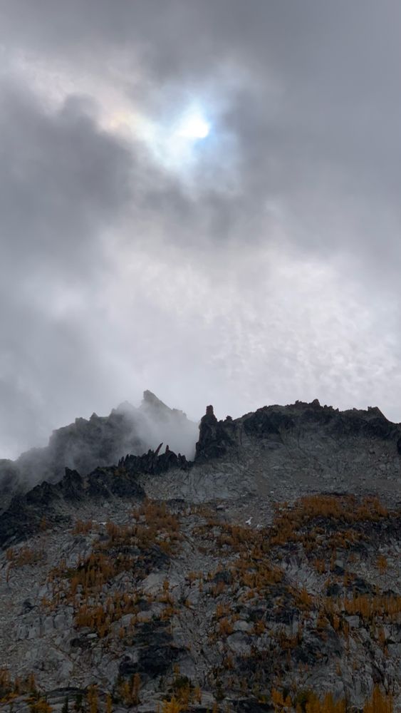 A cloud passes over McClellan Peak at the Alpine Lakes Wilderness in the Enchantments. The blackened granite rock formations look almost like rib bones. Golden larch trees grow along the mountainside.