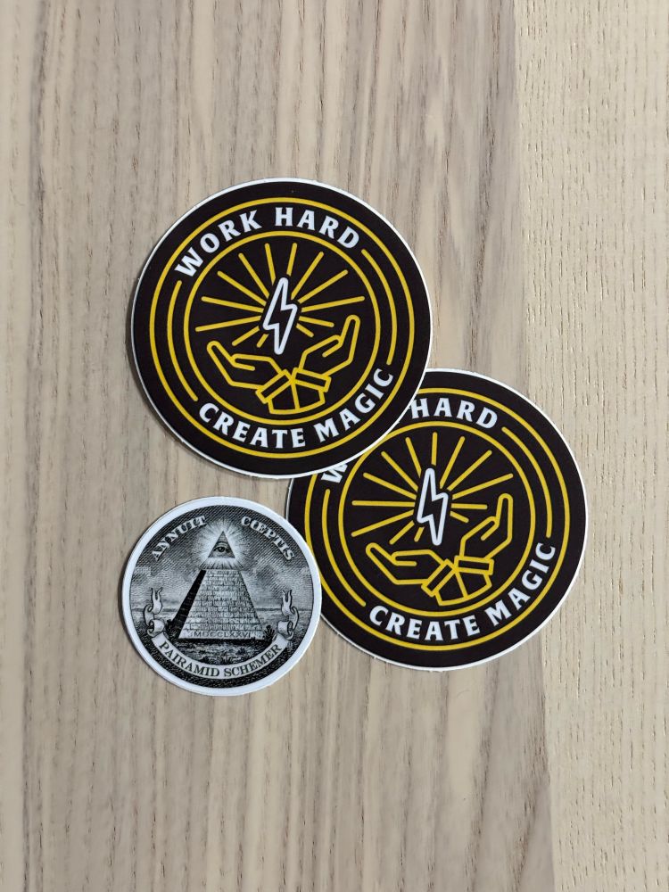 Three stickers on a wooden table, with the captions “Work hard, create magic” and “Pairamid schemer”