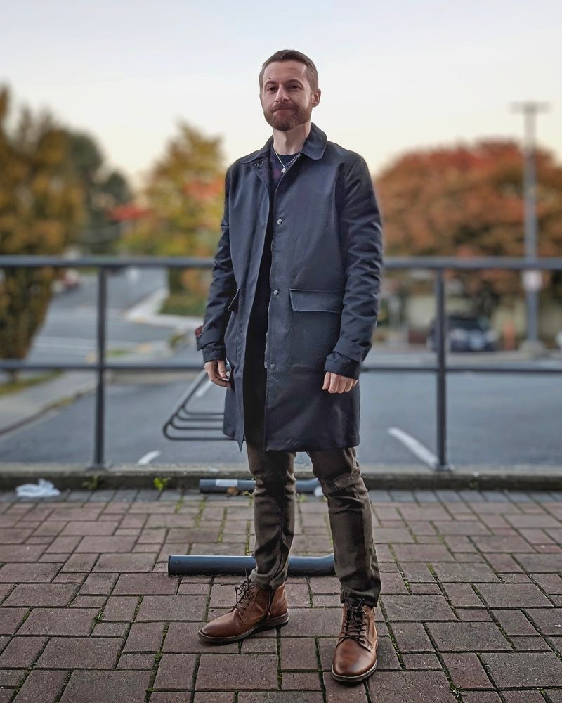 A man stands on a brick sidewalk. They are wearing a long navy jacket. 