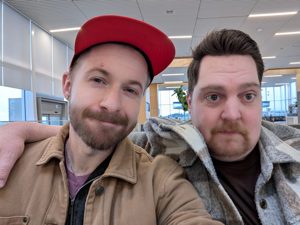 Two men sitting side by side in an airport lounge. One on the left is wearing a red hat. 