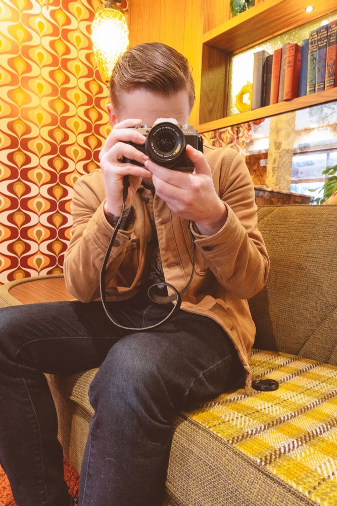 A man sitting on a fabric plaid couch. Surrounding them is 70s inspired living room decor. The man is holding a camera up to their face and pointing straight in front. 