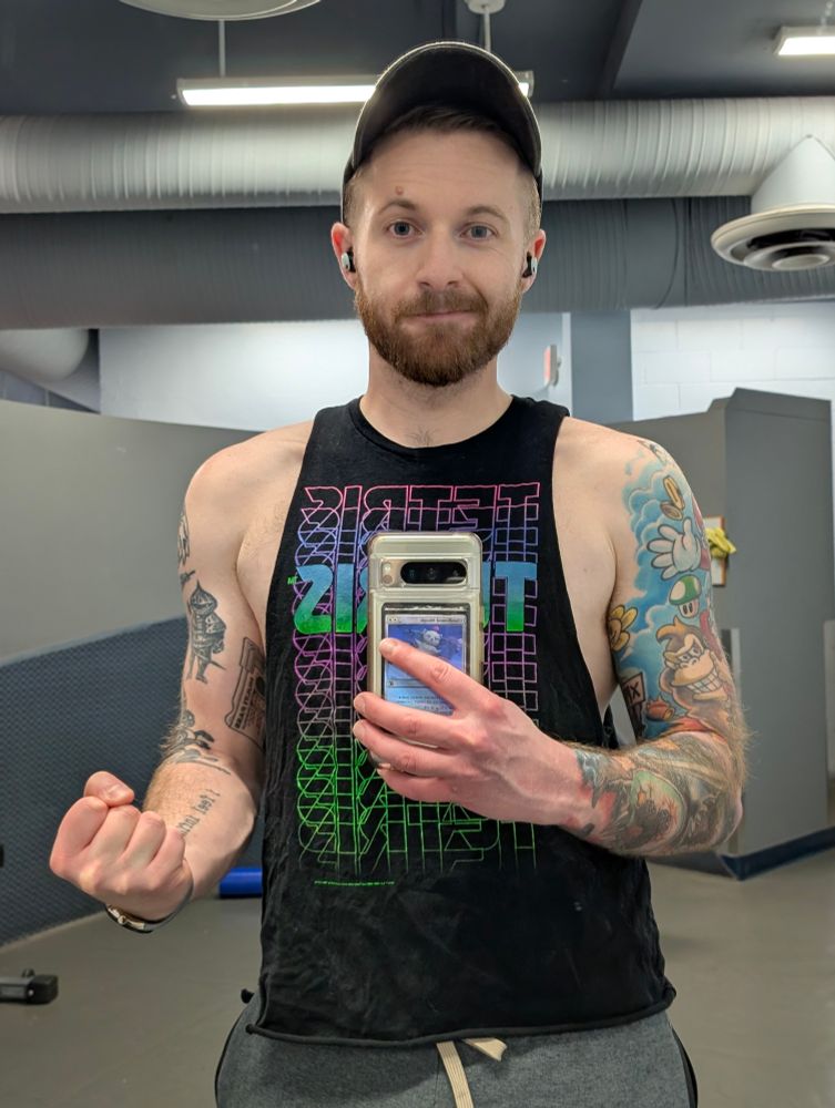 A man flexes his bicep while taking a picture of themself in a gym mirror. 