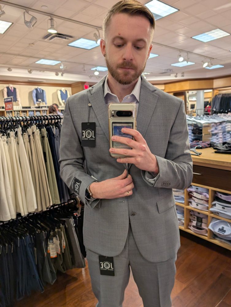 A man taking a picture of themself in a mirror wearing a grey suit with tags attached to it. 