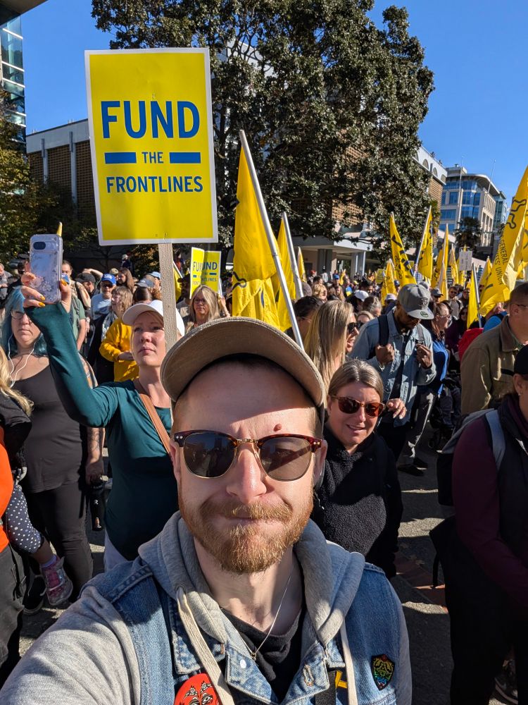 A man standing in a crowd of people holding up signs. One of the signs reads "Fund the Frontlines"