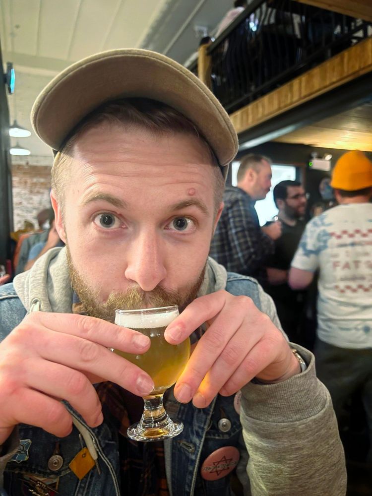 A man wearing a denim jacket looks directly into the camera as they hold a small glass of craft beer up to their mouth. Behind them are patrons in a brewpub. 