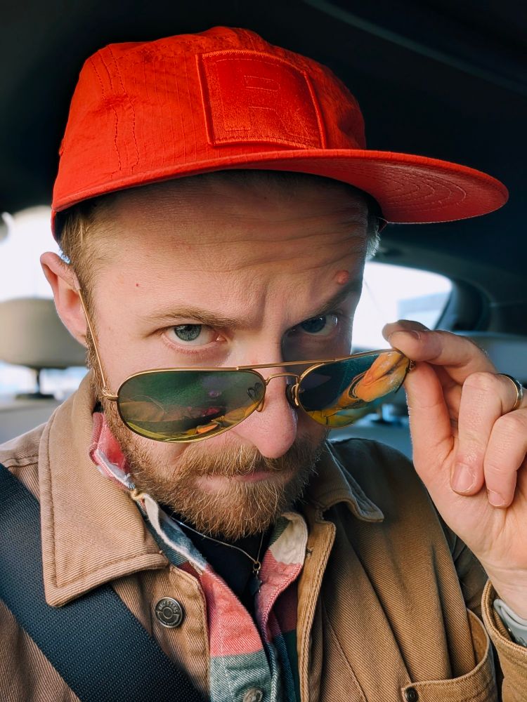 A man peering over their sunglasses in the back of a car. They are wearing a Team Rocket cap and a beige jacket. 