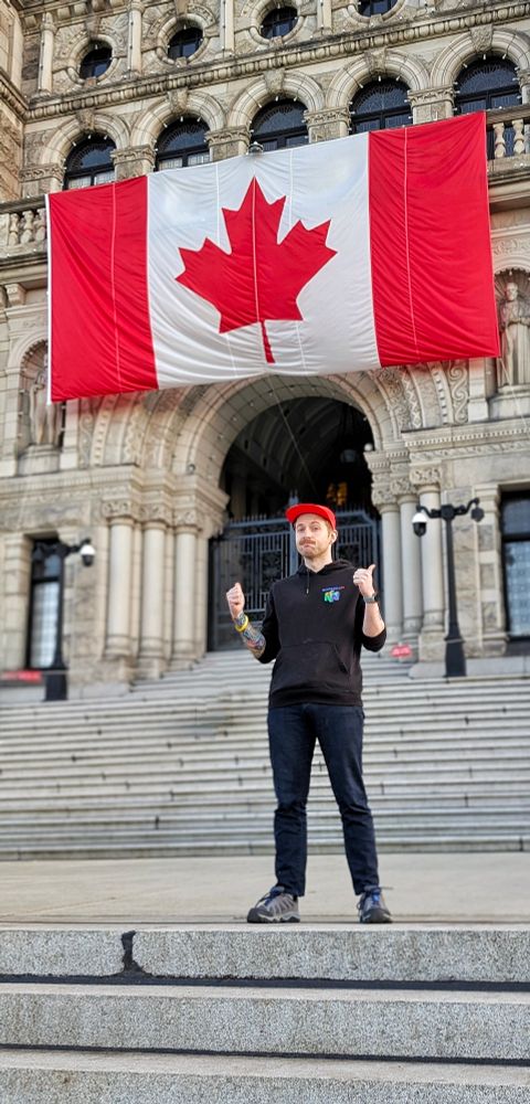 A man standing in front of a parliamentary building with a large Canadian Flag hanging behind them. 