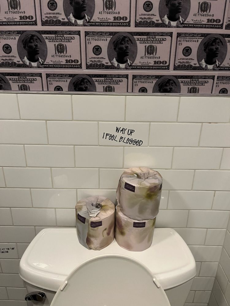 Hip hop style forward wash closet with porcelain decor, branded paper for the derrier, customized tile work featuring culturally iconic quotes and lyrics, along with interior design inspired wallpaper featuring Tupac as president on one hundred dollar bills.