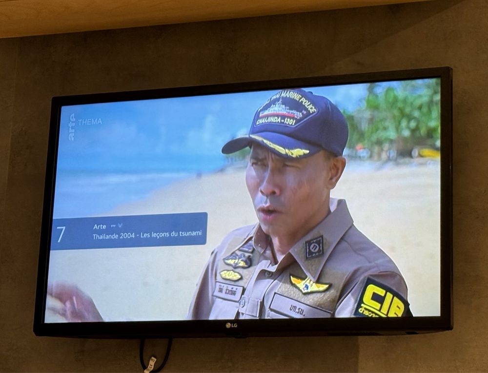 A photo of a hotel tv showing a programme in French about the 2004 Thailand tsunami. The image shows a Thai man in uniform on a beach talking to camera. 