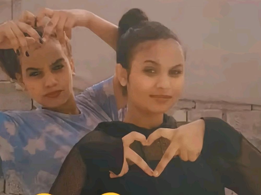 Two young Eritrean posing for a picture, one wearing blue shirt holding her two hands on top of her haid and the other one wearing black making a heart sign with her two fingers. 
