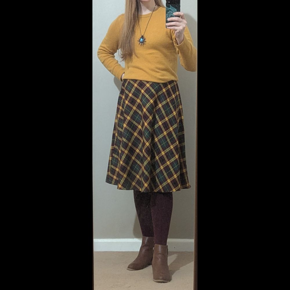 A woman wearing an outfit consisting of a yellow cardigan, labradorite pendant, yellow/green/burgundy plaid skirt, burgundy stockings, and short brown boots.