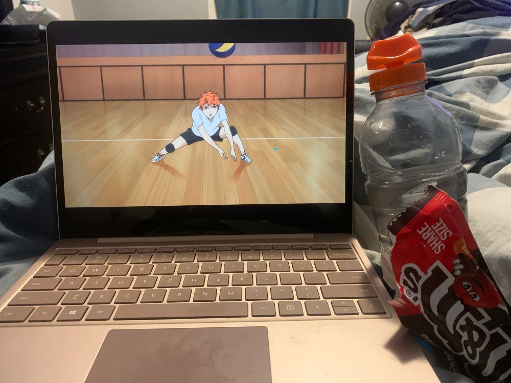 The image consists of a screenshot of the anime Haikyuu on a laptop with the red headed main character Hinata about to return the ball. Next to the laptop sits an opened Gatorade bottle and an open pack of share size milk chocolate M&M’s. 