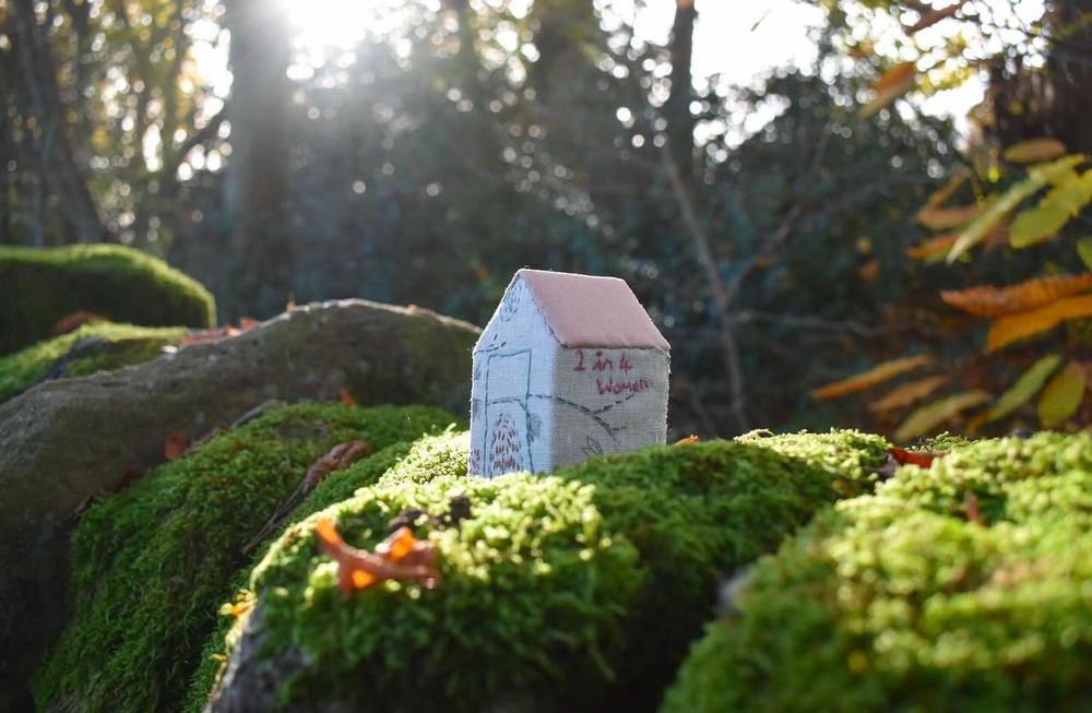 Small textile house sat in the sunlight. It sits on a moss covered wall with trees in the background. 