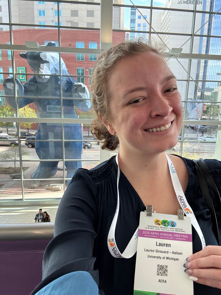 Lauren holds up a badge with her name and the AERA logo of a yellow, green, and blue circle. There is a large blue bear behind her. 