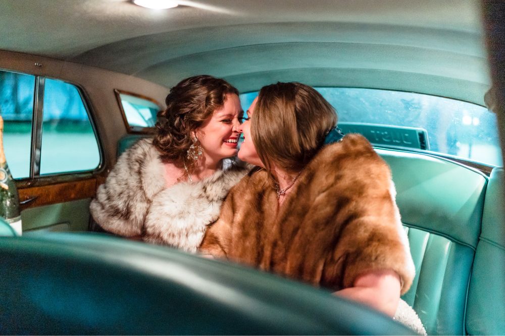 Lauren and her wife Maddie in a vintage car with teal seats smile and laugh together.