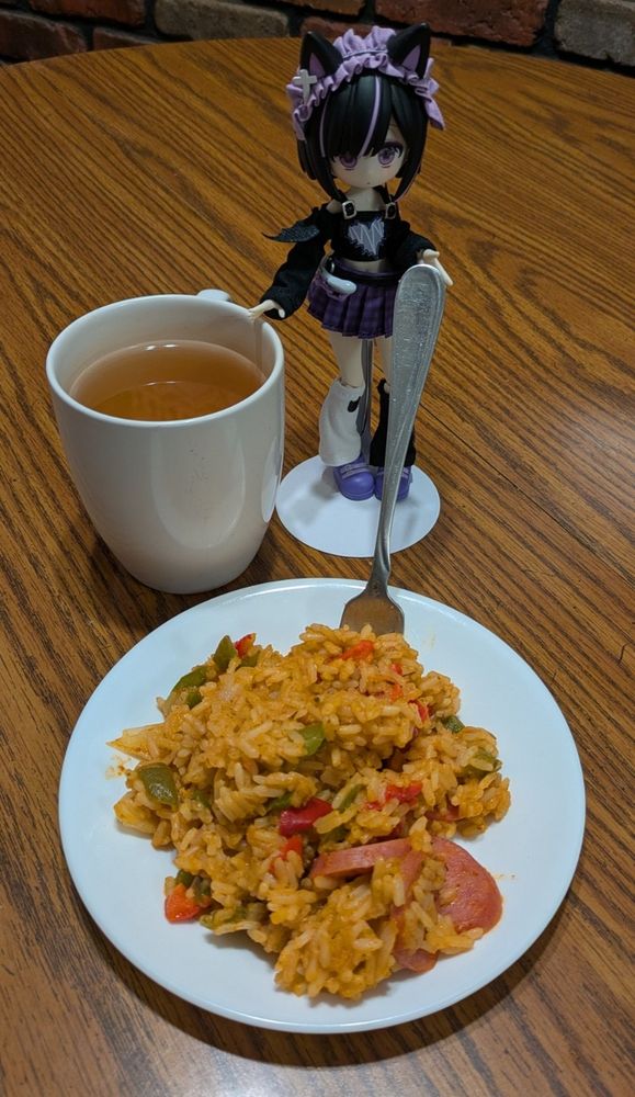 A small doll wearing purple clothes and a skirt holds a fork. A plate has a helping of Cajun jambalaya, which is rice, peppers, and Italian sausage. A cup of chamomile tea sits next to the plate and doll.
