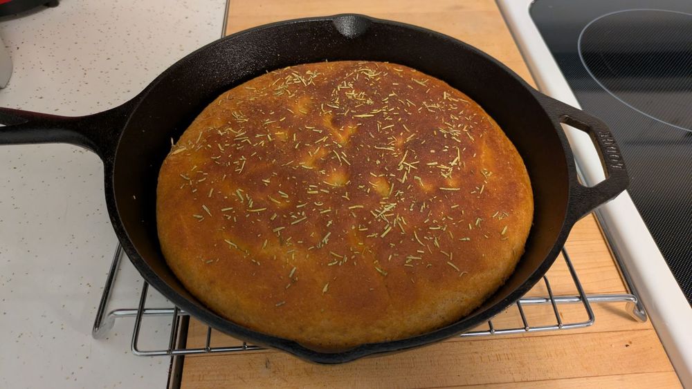 The pan bread has been cooked and is sitting on a wire rack, still in a black cast iron pan, to cool. The bread is somewhat dark on top, but is not burned.