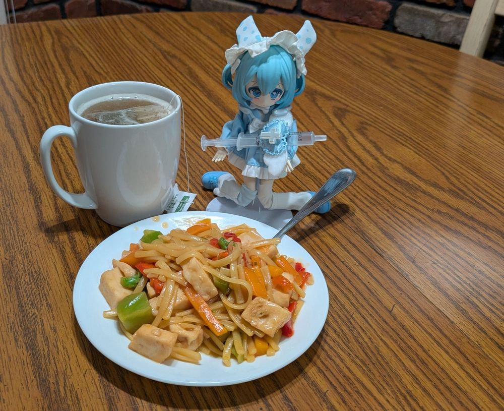 A small doll dressed in a light blue nurse outfit and holding a toy syringe kneels next to a plate of chicken stir fry and a cup of green tea. There is a tea bag still in the cup.