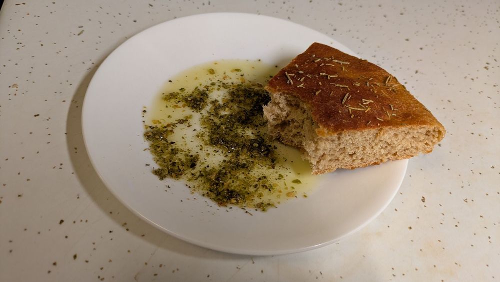 A wedge of pan bread sits on a white ceramic plate. There is a mix of olive oil, various herbs, and cheese on the plate. The bread has been dipped into the mix of oil and a bite has been taken out of it.
