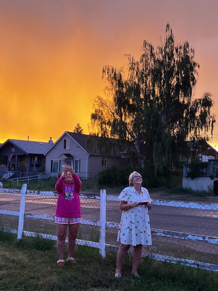 Two women stand in a yard, smiling up at the orange sunset overhead.